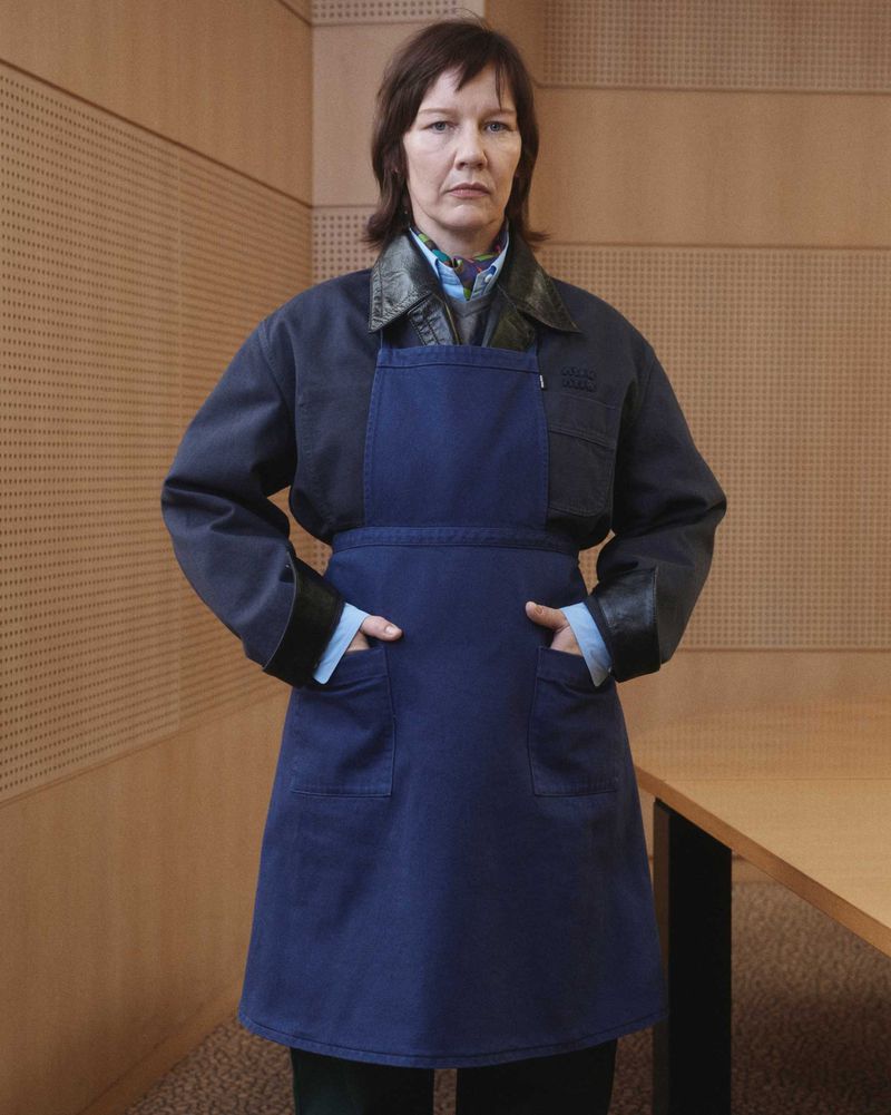 An adult woman with medium-length brown hair wears an apron over a jacket with leather trim, standing indoors in front of a paneled wall.