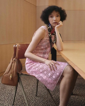 Model wearing pink lace dress seated at a wooden table with brown handbag hanging on chair