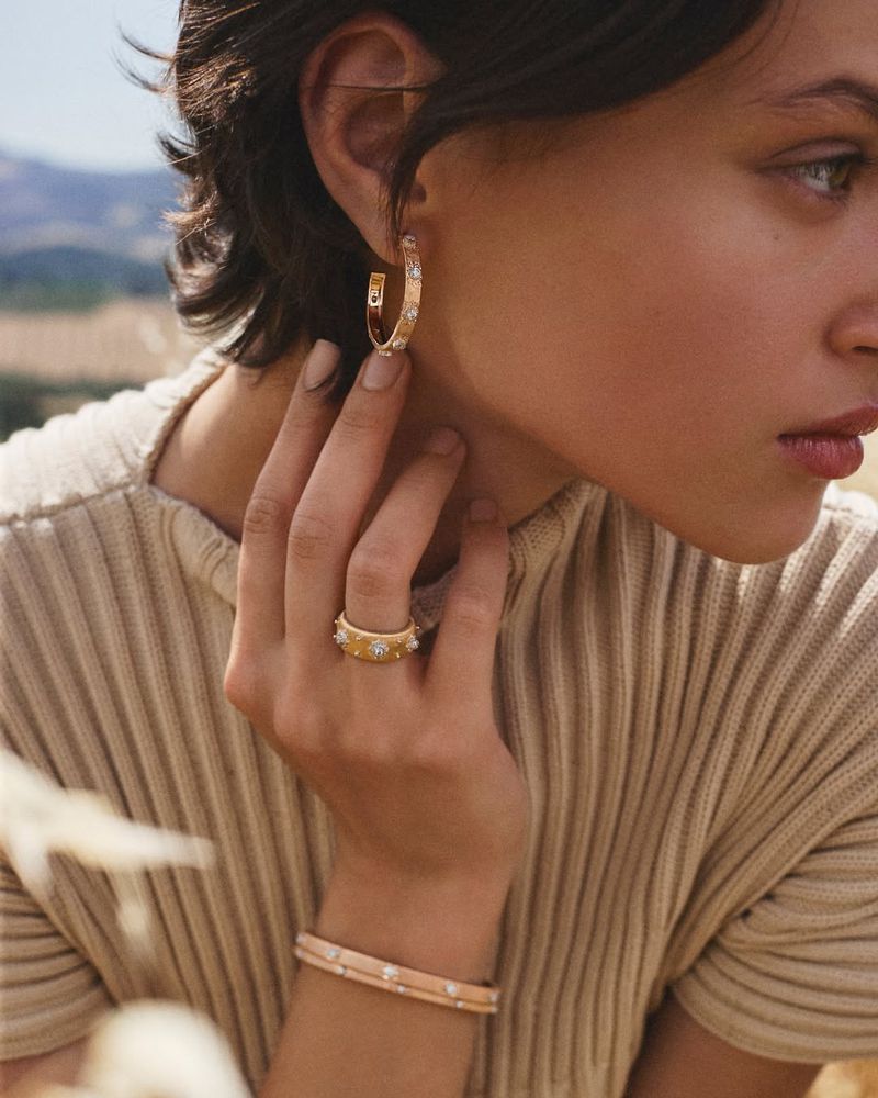 Woman's profile showcasing gold hoop earrings and a ring with diamond accents against a blurred outdoor background with wheat field.