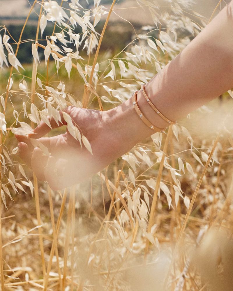 Close-up of a woman's hand with bracelets gently touching wheat stalks in a sunlit field, highlighting jewelry and nature.