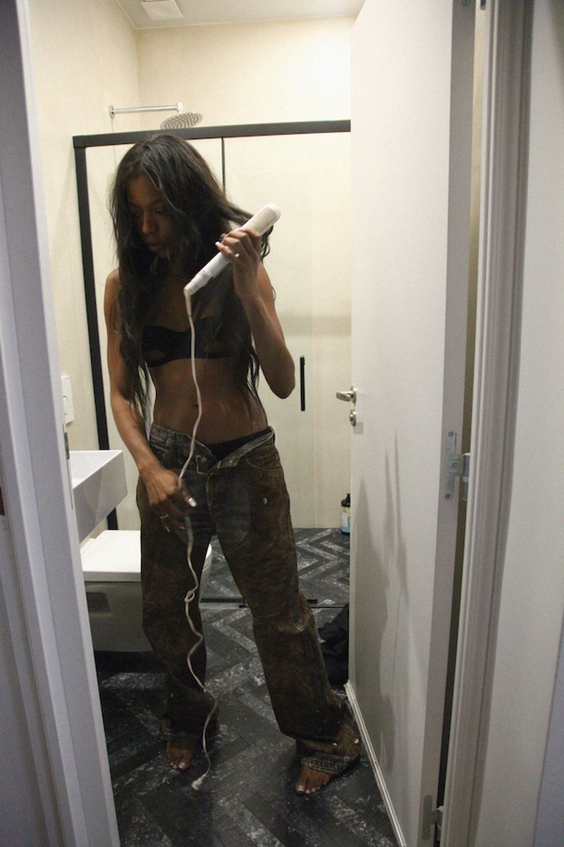 Woman with long dark hair in casual clothing, using a hair styling tool inside a brightly lit bathroom with white fixtures.