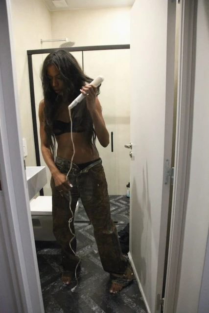 Woman with long dark hair in casual clothing, using a hair styling tool inside a brightly lit bathroom with white fixtures.
