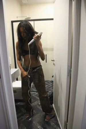 Woman with long dark hair in casual clothing, using a hair styling tool inside a brightly lit bathroom with white fixtures.