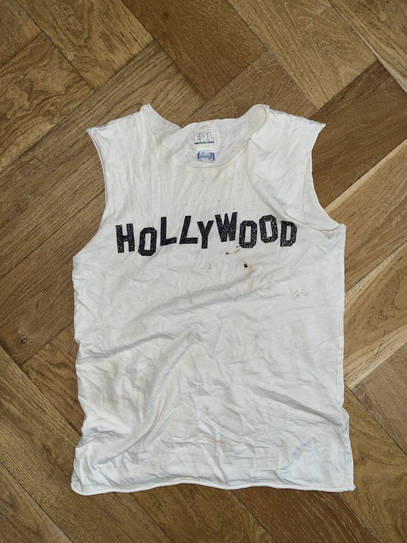 White sleeveless t-shirt with black 'Hollywood' text displayed on a herringbone wood background. Vintage fashion top.
