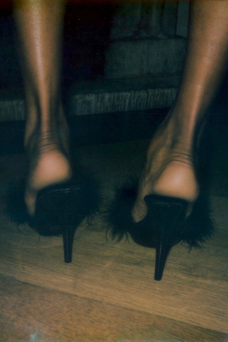 Close-up, low-angle shot of an adult female's bare legs and feet wearing black, feathered high-heeled shoes, standing on a wooden surface.