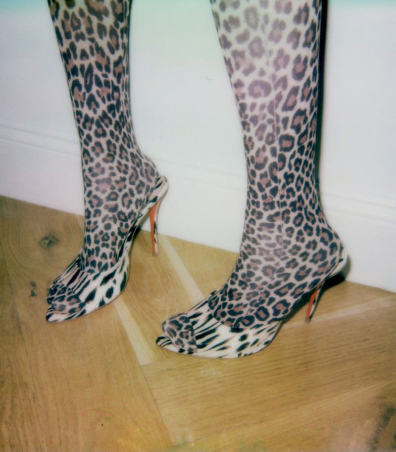 Woman's legs with leopard print tights and matching open-toe high heel shoes standing on a wood floor with a white wall background.