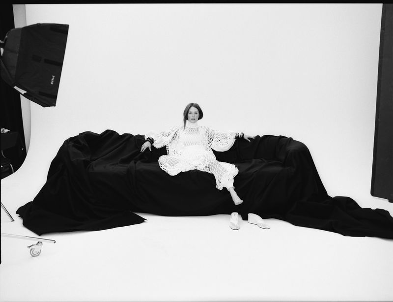 An adult woman sits on a black couch wearing a white crocheted outfit in a photo studio, with a light and white backdrop.