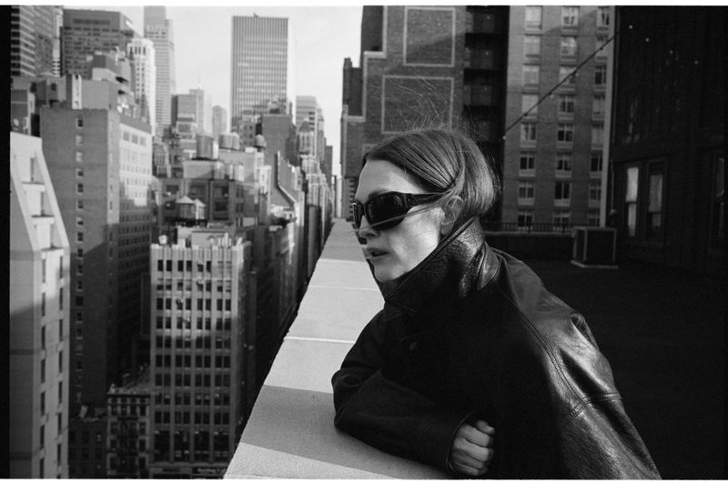 Adult female with sunglasses leans on a rooftop ledge overlooking a dense urban skyline in a black-and-white photograph.