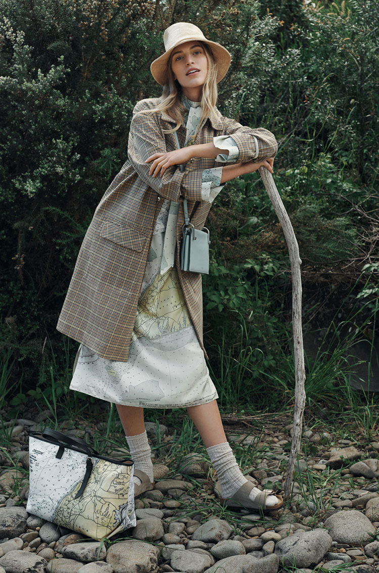 Woman in plaid coat and sunhat leans on walking stick in a forest setting wearing sandals and socks carrying a tote bag.