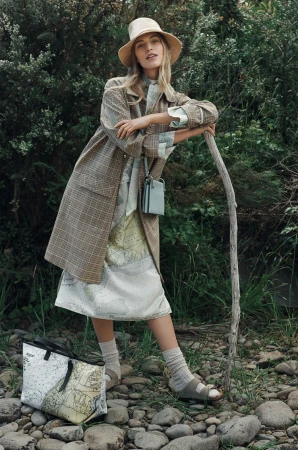 Woman in plaid coat and sunhat leans on walking stick in a forest setting wearing sandals and socks carrying a tote bag.