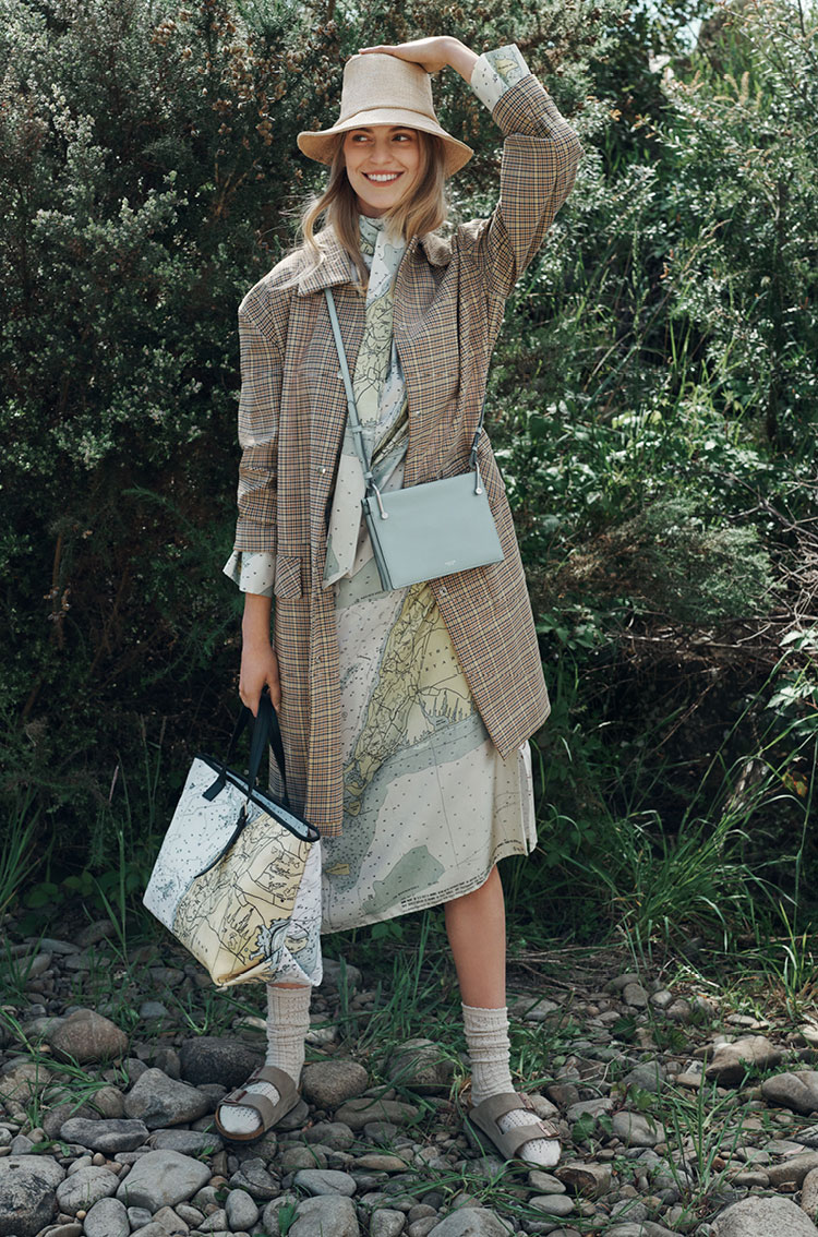 Full-shot of a woman in a hat, patterned dress, and coat, carrying a tote bag and crossbody bag, standing outdoors in a rocky area.