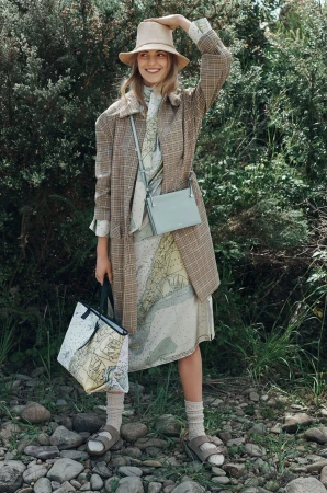 Full-shot of a woman in a hat, patterned dress, and coat, carrying a tote bag and crossbody bag, standing outdoors in a rocky area.