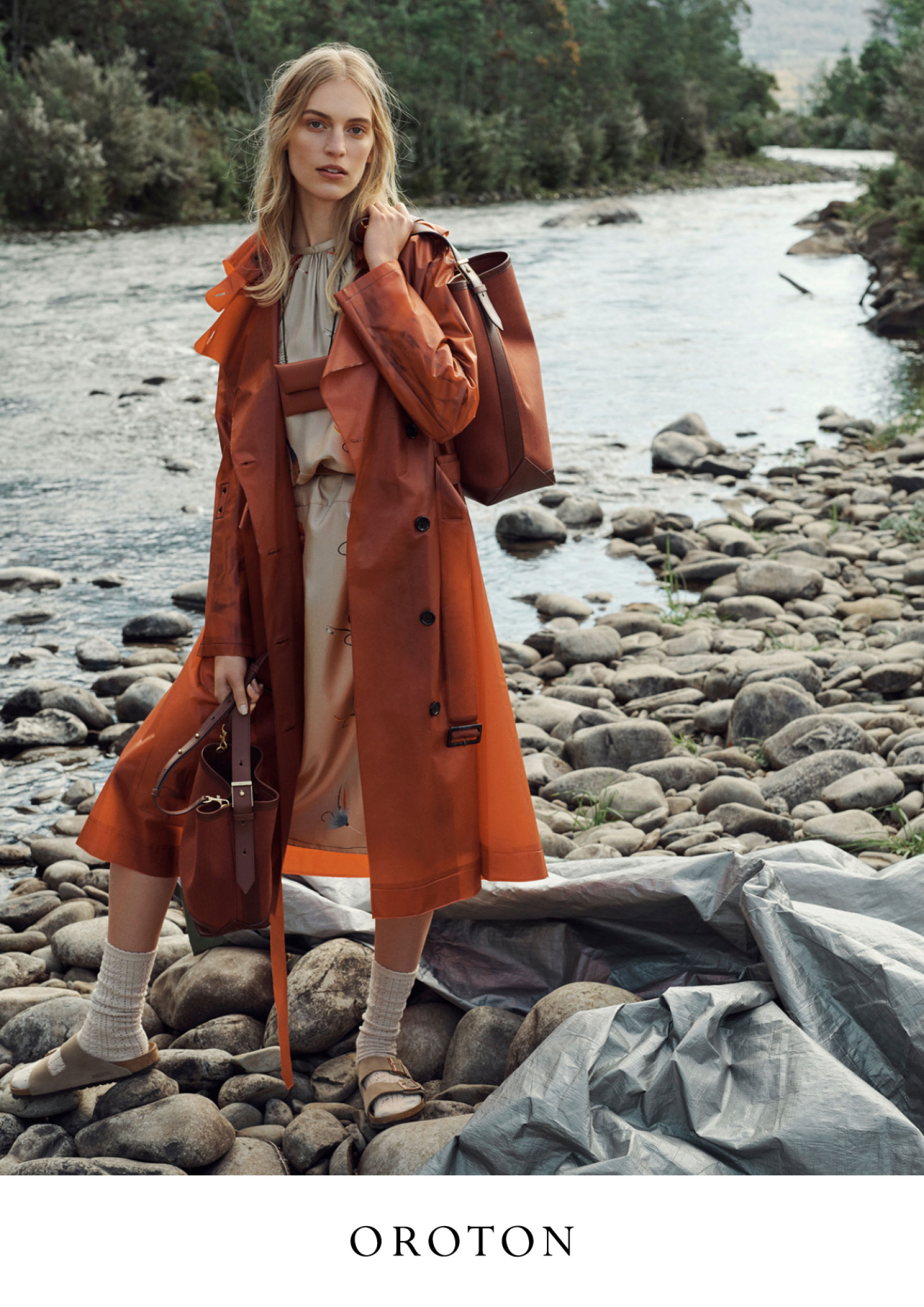 Fashion editorial shot of a blonde woman in a trench coat holding purses standing on rocks by a river in an outdoor setting.