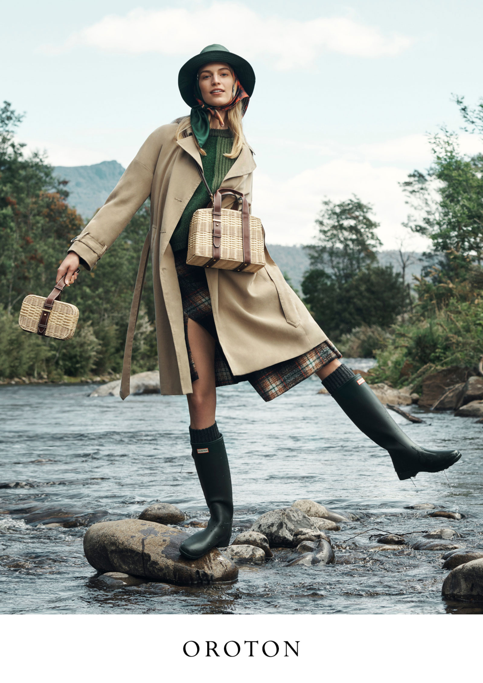Fashion image of a woman in a beige trench coat, plaid skirt, and rain boots, standing on rocks in a river, carrying wicker bags.