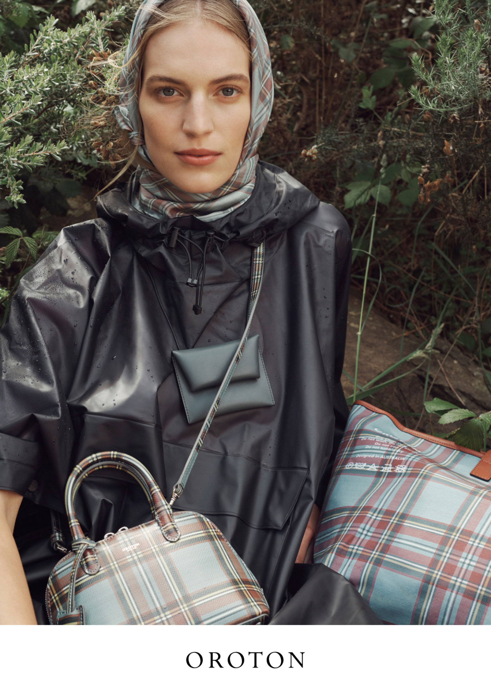 Woman in a hooded raincoat, scarf, and plaid handbags poses outdoors with green foliage in the background, showcasing fashion accessories.