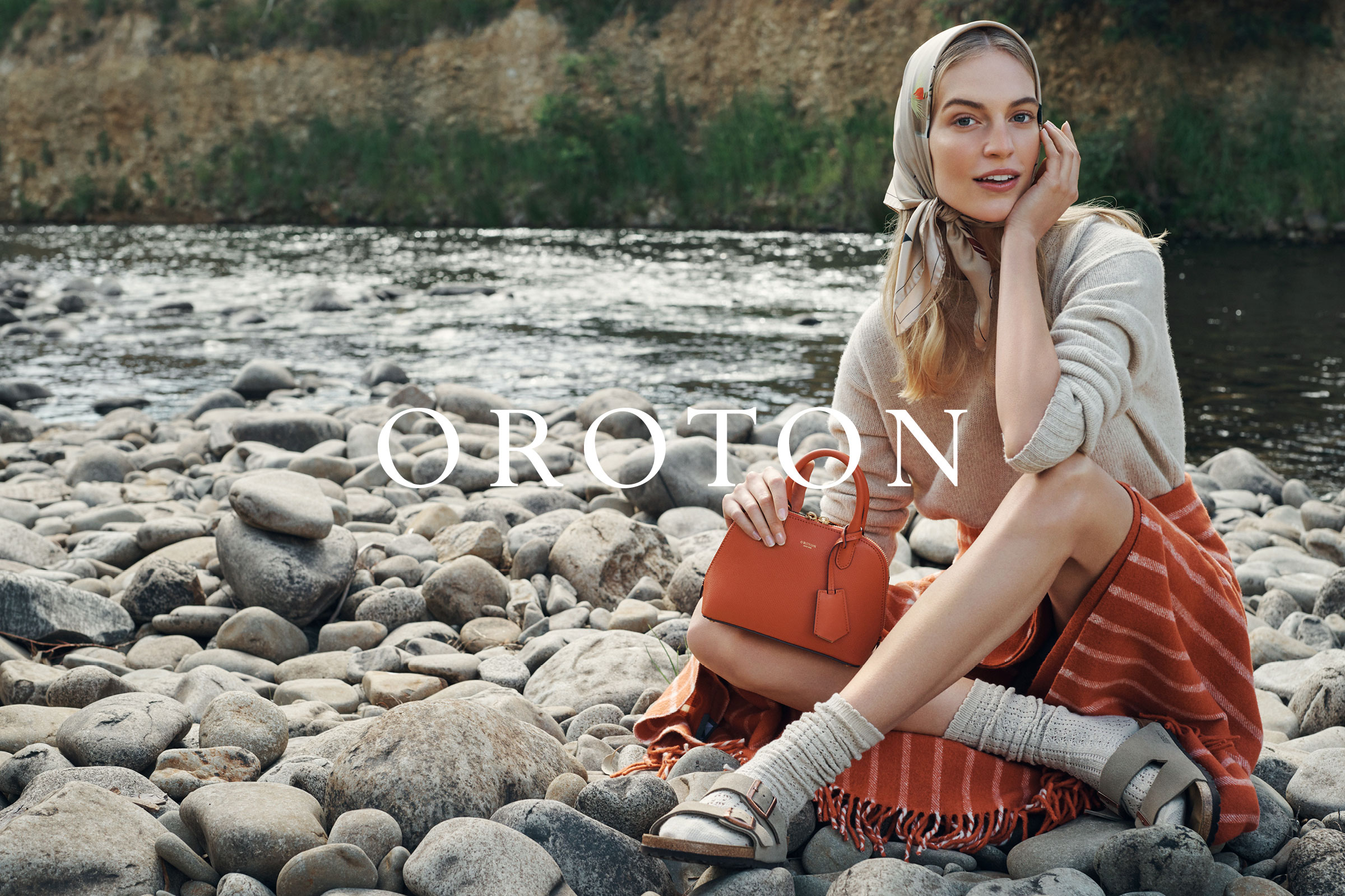 Blonde woman poses by a river with gray rocks, wearing a headscarf, sweater, skirt, and sandals, holding an orange Oroton handbag.