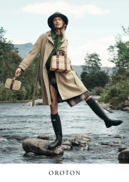 Model wearing a trench coat and tall green boots standing on rocks in a shallow river, holding woven wicker handbags.