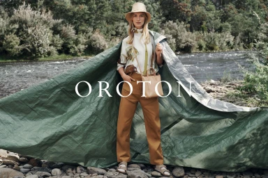 Model Vanessa Axente wearing a tan hat and trousers with a tarp draped behind her by a river for Oroton Winter.