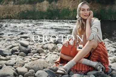 Model seated on a rocky riverbank wearing a cream sweater, patterned headscarf, and orange leather handbag for Oroton Winter campaign