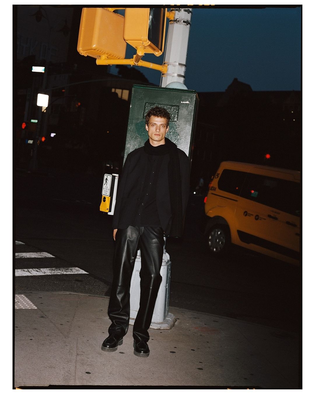 Male fashion model poses on a city street at night near a crosswalk signal, wearing leather pants, a dark shirt and dark shoes.