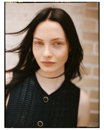 Portrait of a woman with dark hair and a black button-down top against a light textured brick background.