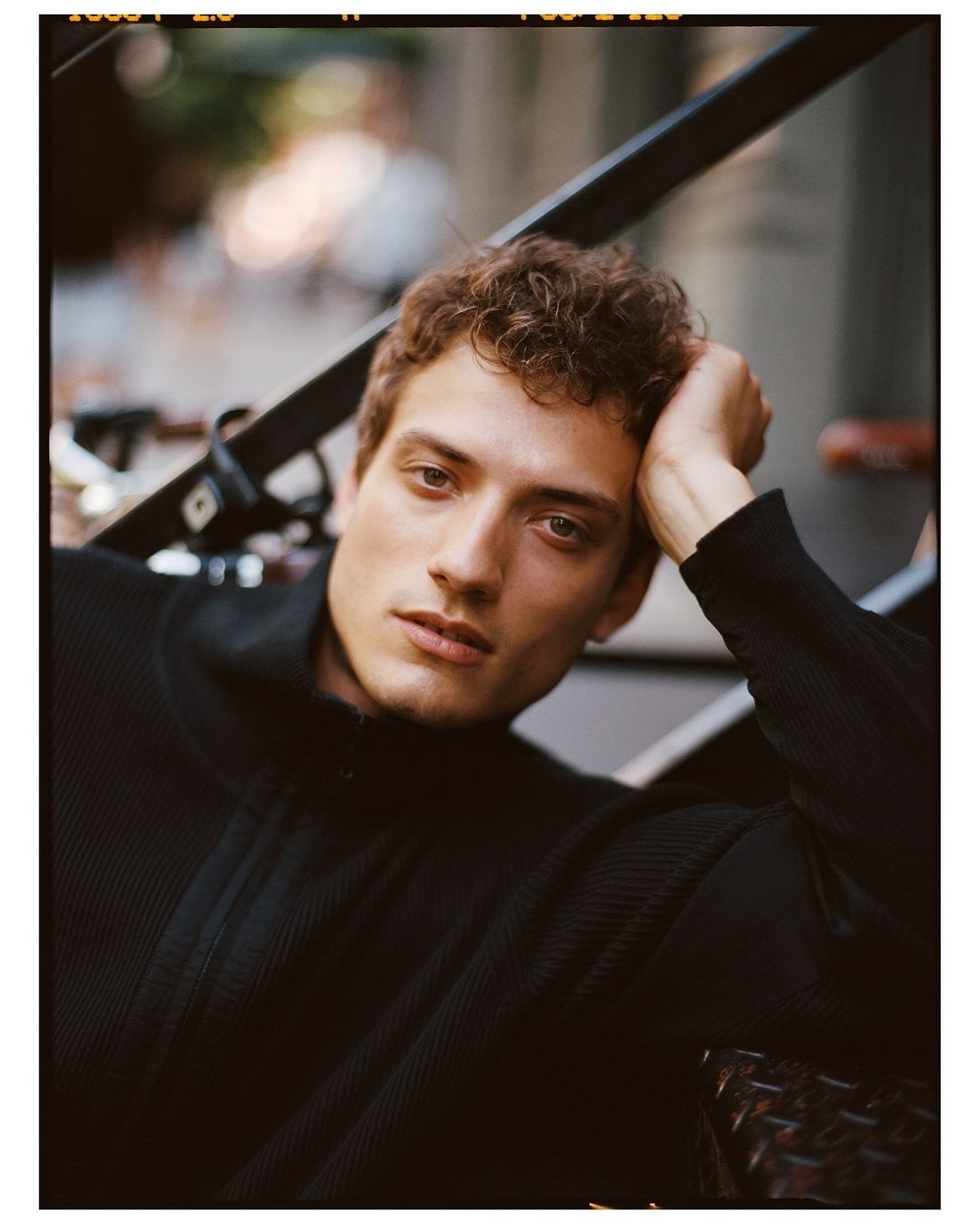 Indoor portrait of a fair-skinned adult male with curly hair wearing a black ribbed sweater, posing on metal stairs looking at camera.
