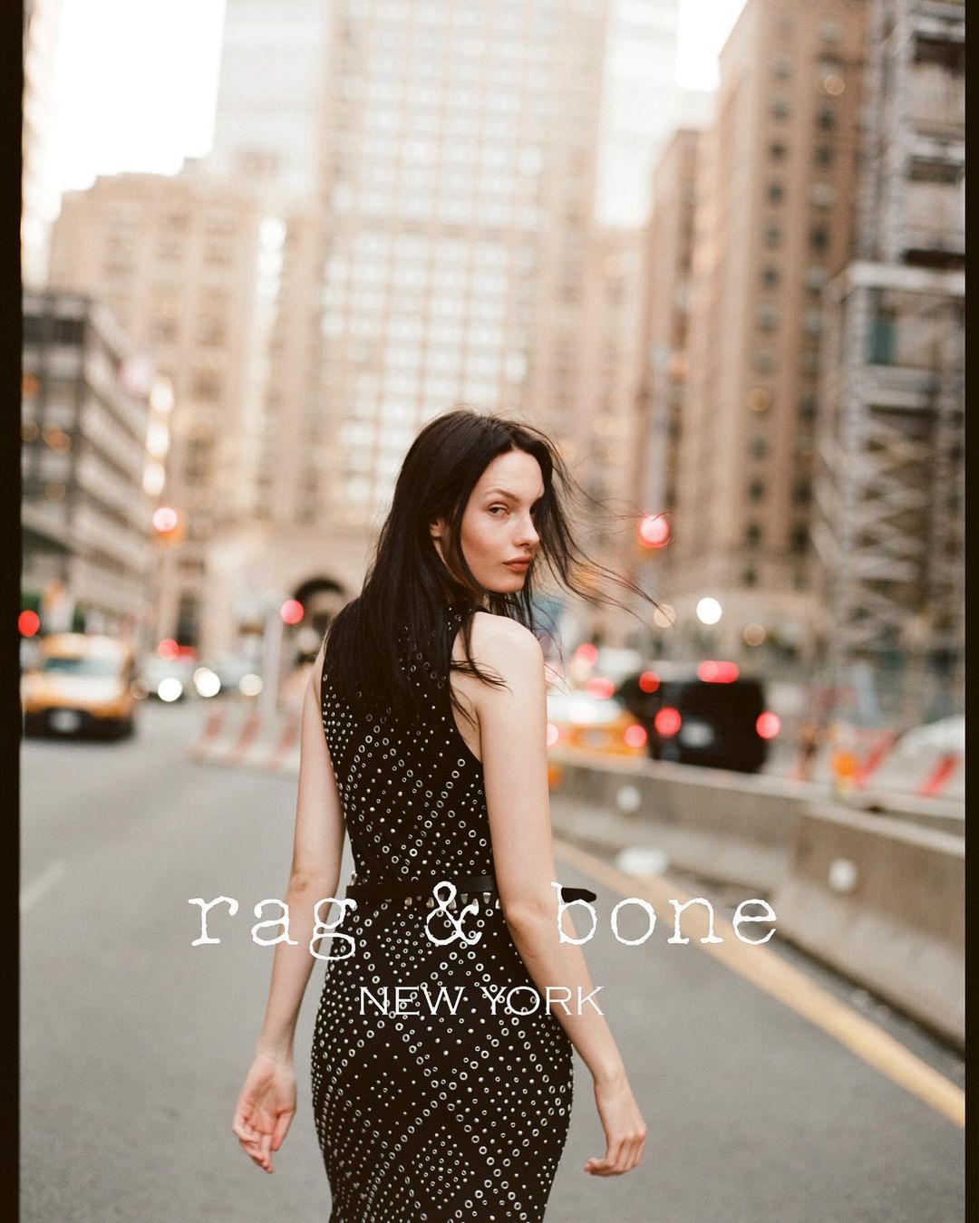 A woman in a black dress with silver accents walks down a city street in New York City, with cars and buildings in the background.