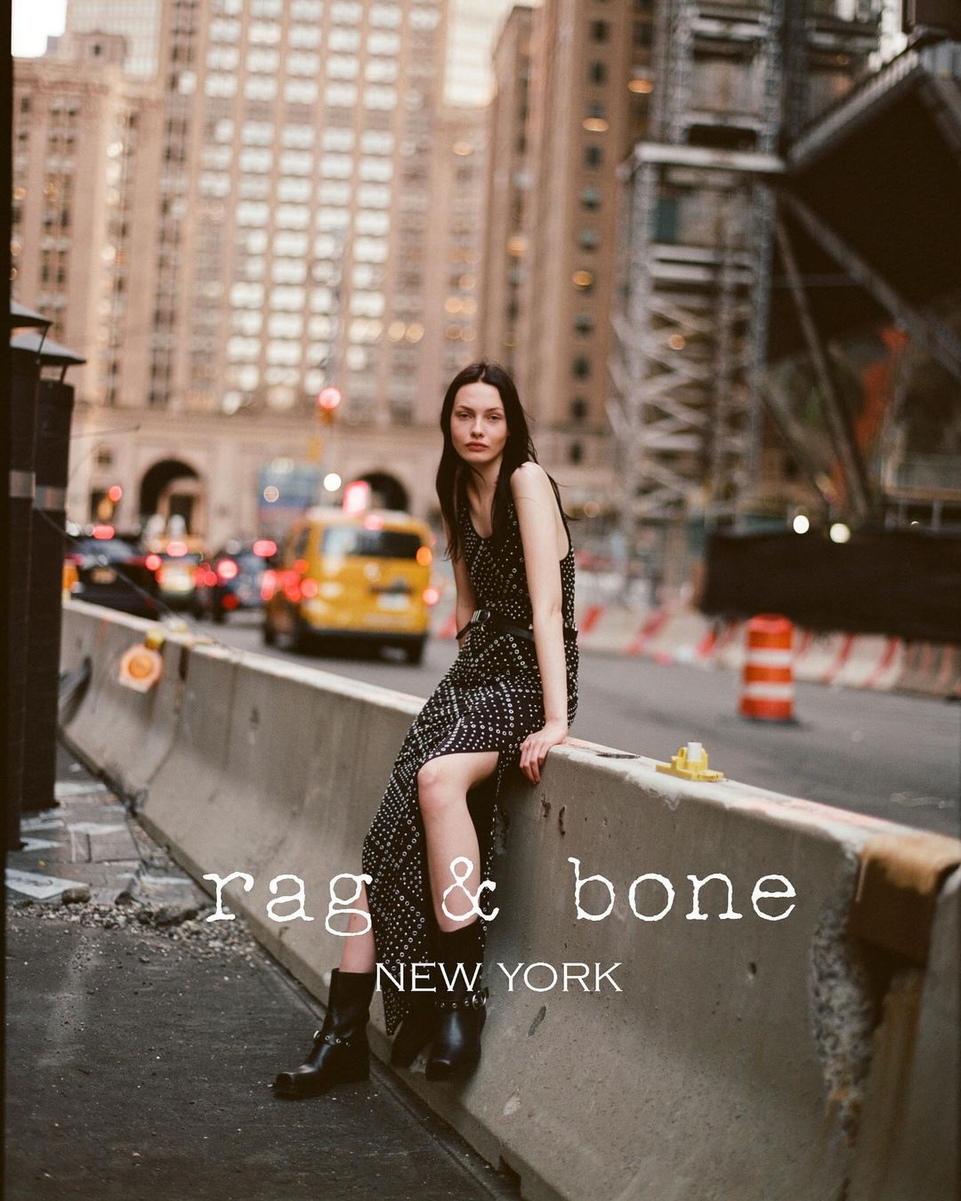 Fashion shot featuring a female model in a polka dot dress and boots, posing on a concrete barrier in an urban setting with city buildings.