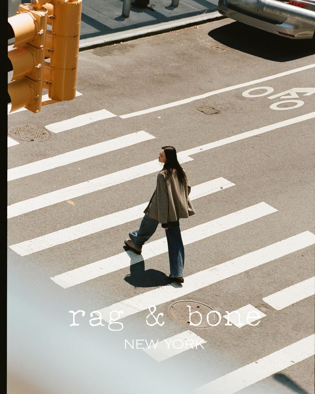 A woman in a coat and jeans walks across a pedestrian crosswalk in an urban setting with a traffic light and building in the background.