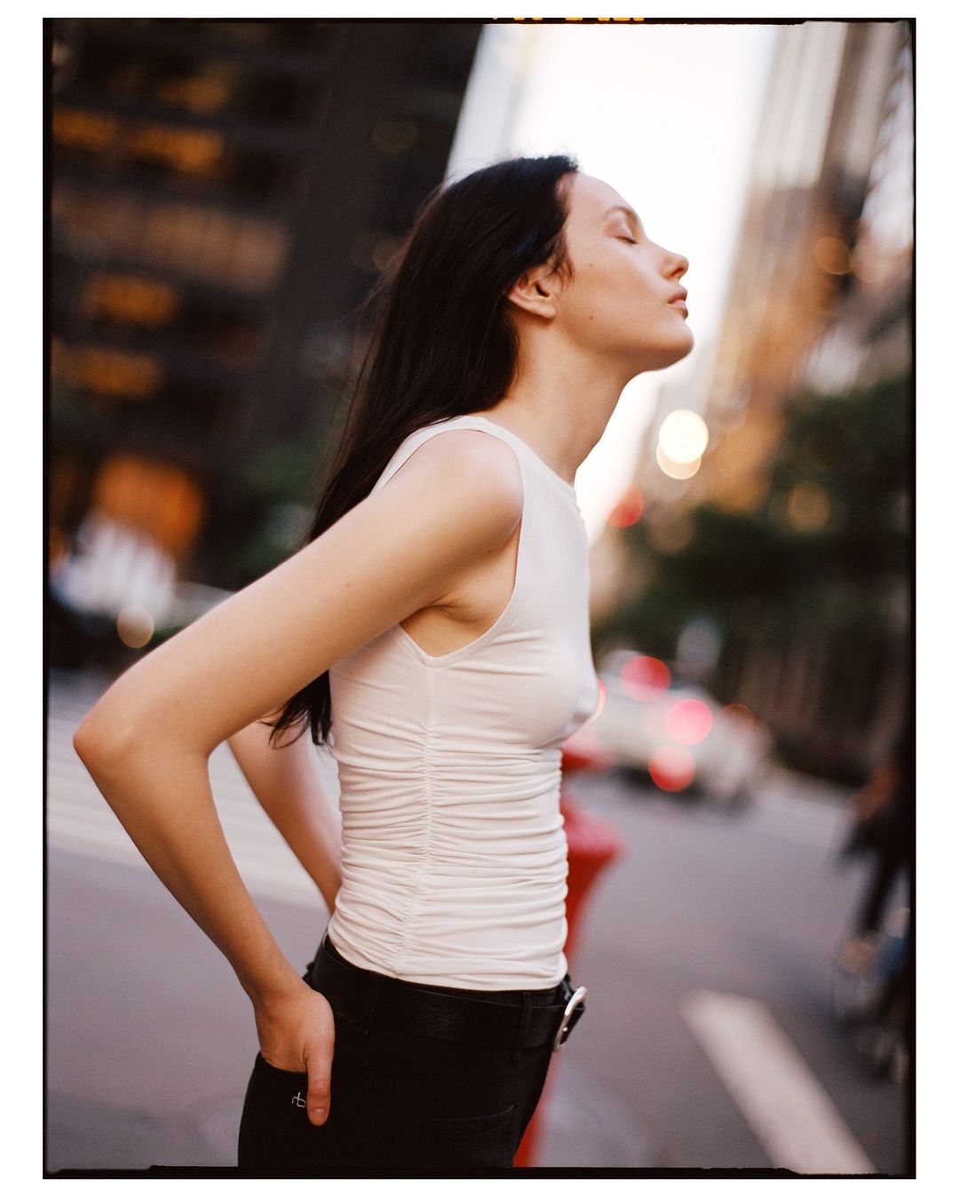 Woman in sleeveless top stands in city street with eyes closed and head tilted back, illuminated by natural light, with buildings in background.
