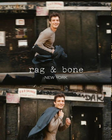 Fashion shot of a young man smiling and walking in an urban setting, wearing a sweater and carrying a coat over his shoulder. 