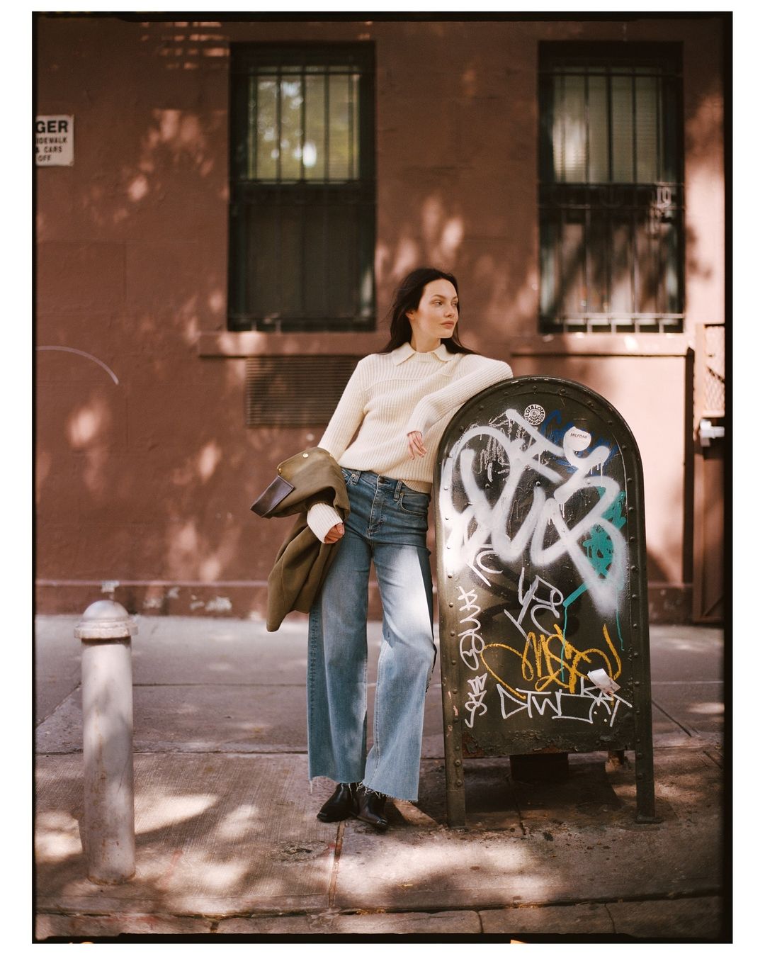 A woman with long dark hair stands on a city sidewalk in a cream sweater and jeans leaning against a graffiti-covered mailbox.