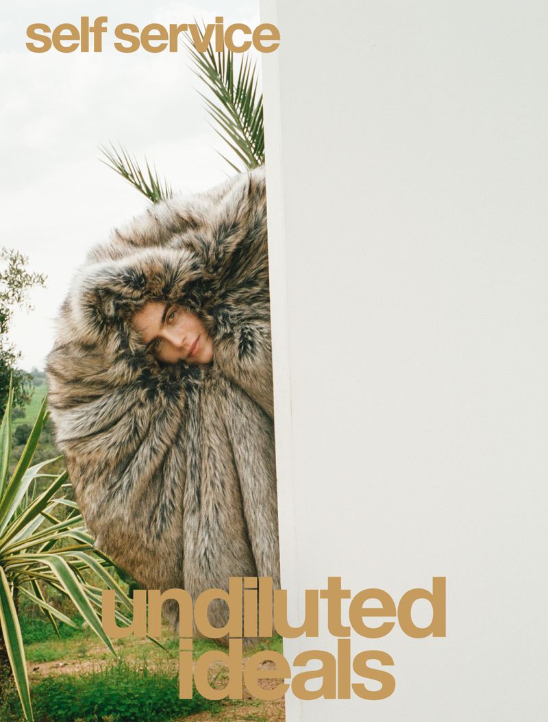 Model wearing a voluminous fluffy faux fur coat peeking from behind a white architectural wall outdoors