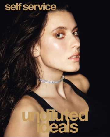 Portrait of a woman with dark hair and a diamond choker on a black background with text overlay from Self Service magazine cover.