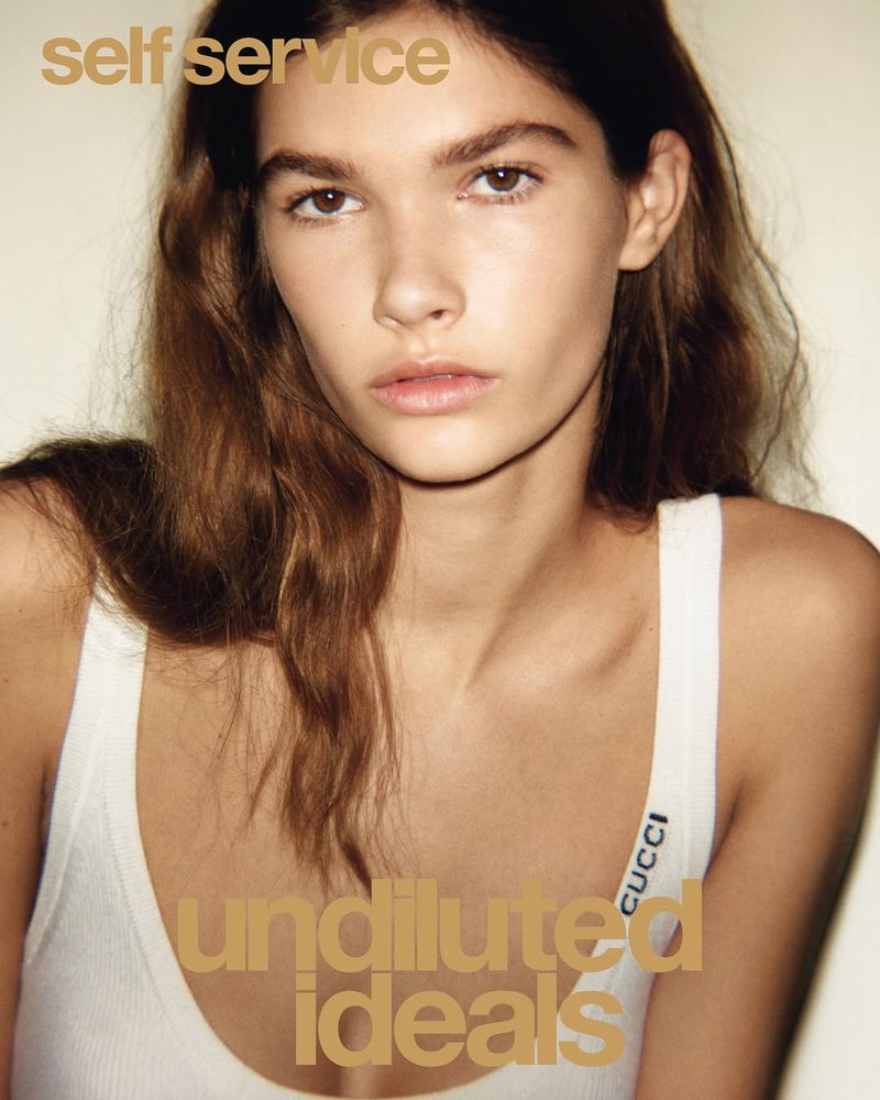 Portrait of a young fair-skinned woman with wavy brown hair wearing a white tank top, posing against a neutral backdrop, lit with soft lighting.