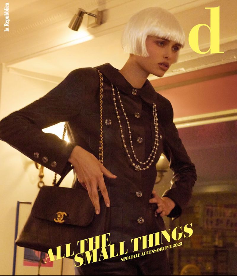 Fashion editorial shot of a model in a dark jacket with a pearl necklace, holding a handbag, against a neutral indoor backdrop.