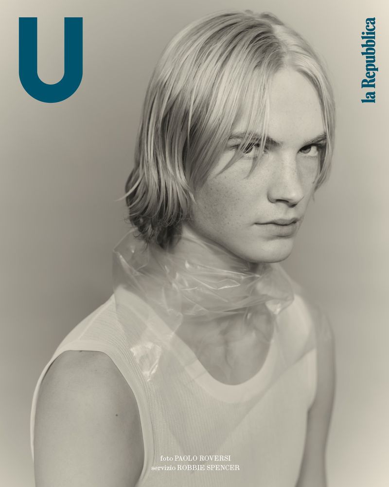 Portrait of a male model with light blonde hair wearing a white top and clear plastic collar, against a neutral background in a studio setting.