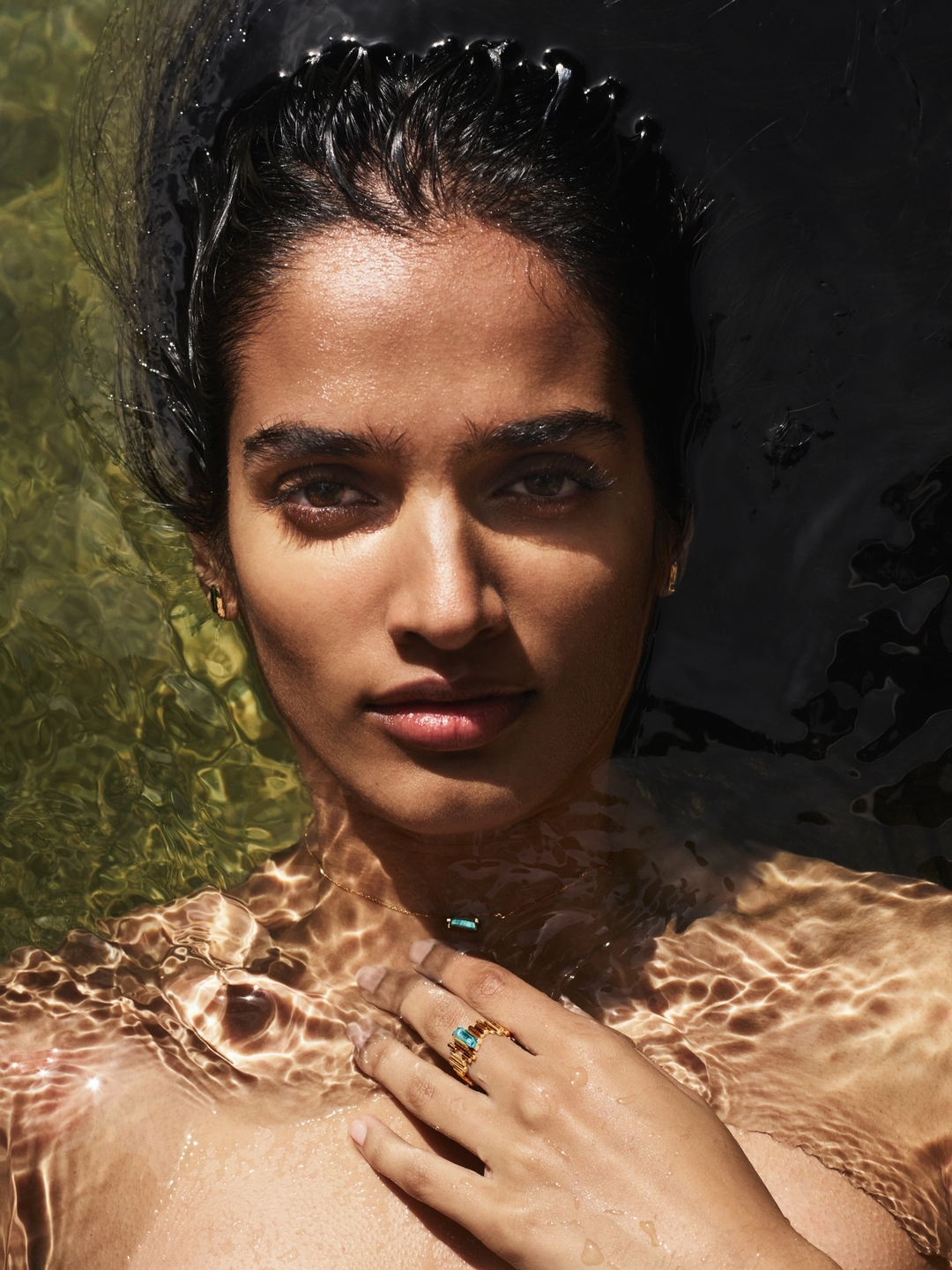 Close-up shot of a woman with wet dark hair, gold ring, and fair skin in shallow water. One adult female with arms around herself.