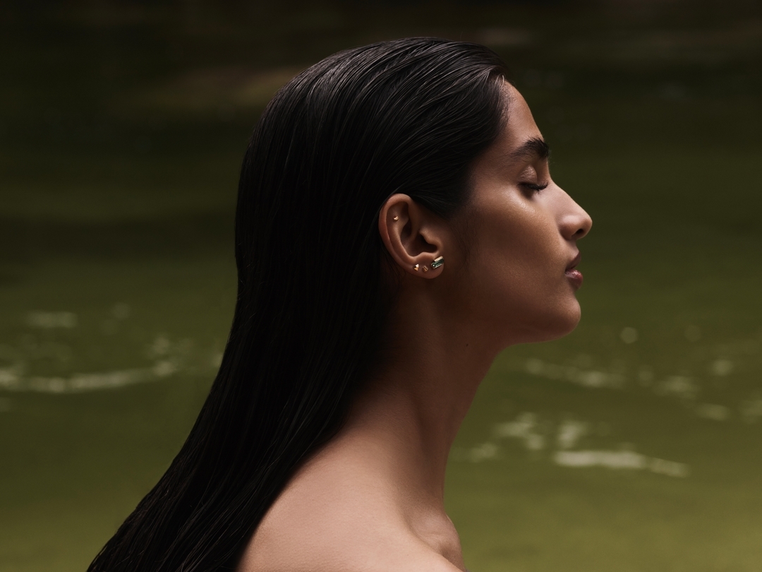 Profile of an adult woman with bare shoulders, dark hair, and gold earrings against a blurred green outdoor background, natural lighting.