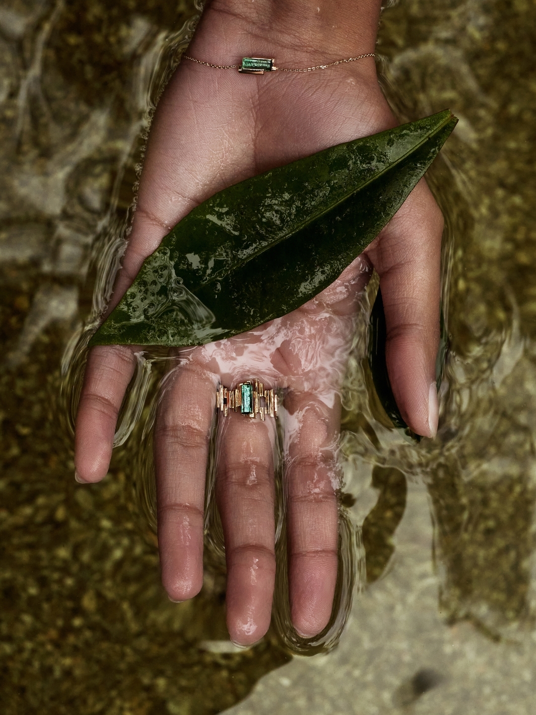 Hand holding a ring and leaf in water. The ring has a green stone and a gold band. A bracelet with a green stone is on the wrist.