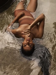 Model lying in shallow water wearing brown swimwear and delicate gold jewelry for Sarah & Sebastian