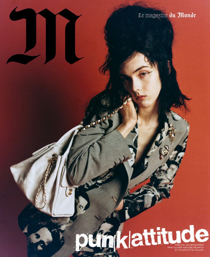 Model with dark beehive hair wearing a patterned blazer and carrying a white handbag against a red background.