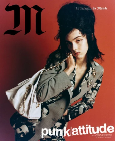 Model with dark beehive hair wearing a patterned blazer and carrying a white handbag against a red background.