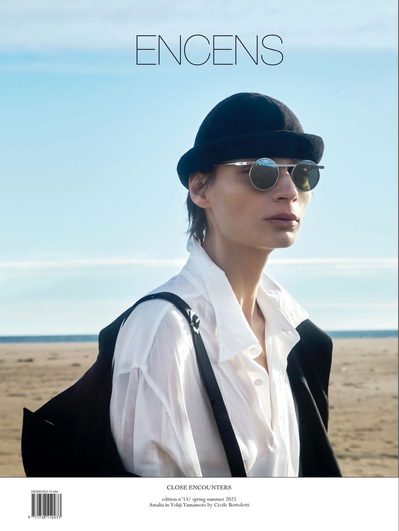 Fashion editorial portrait of a woman in a hat and sunglasses on a beach, promoting Encens magazine, evoking a sense of coastal style.