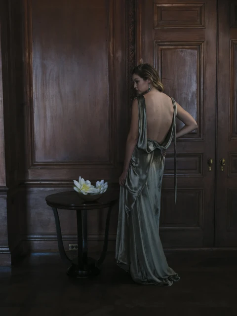 Woman in an elegant dress poses near a table with a decorative white flower, creating a sophisticated and formal indoor ambiance.