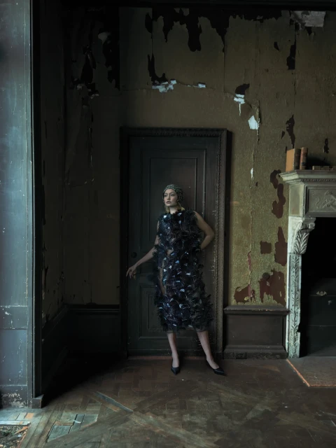 Fashionable woman in feather dress stands in dilapidated room with peeling paint and boarded-up doorway, creating a haunting, editorial image.