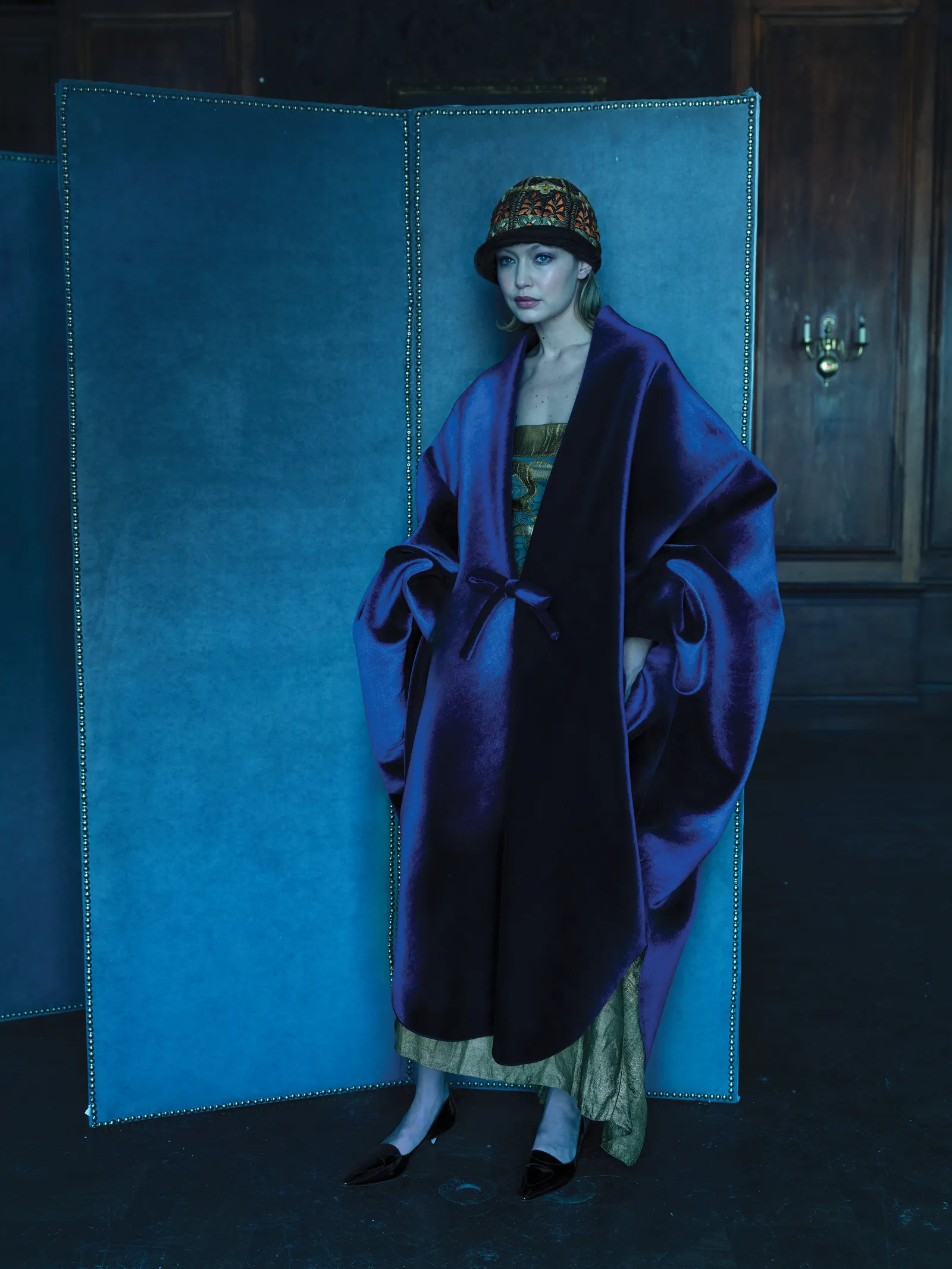 Woman in oversized purple coat with decorated cloche hat stands in front of a blue folding screen in a formal interior setting.