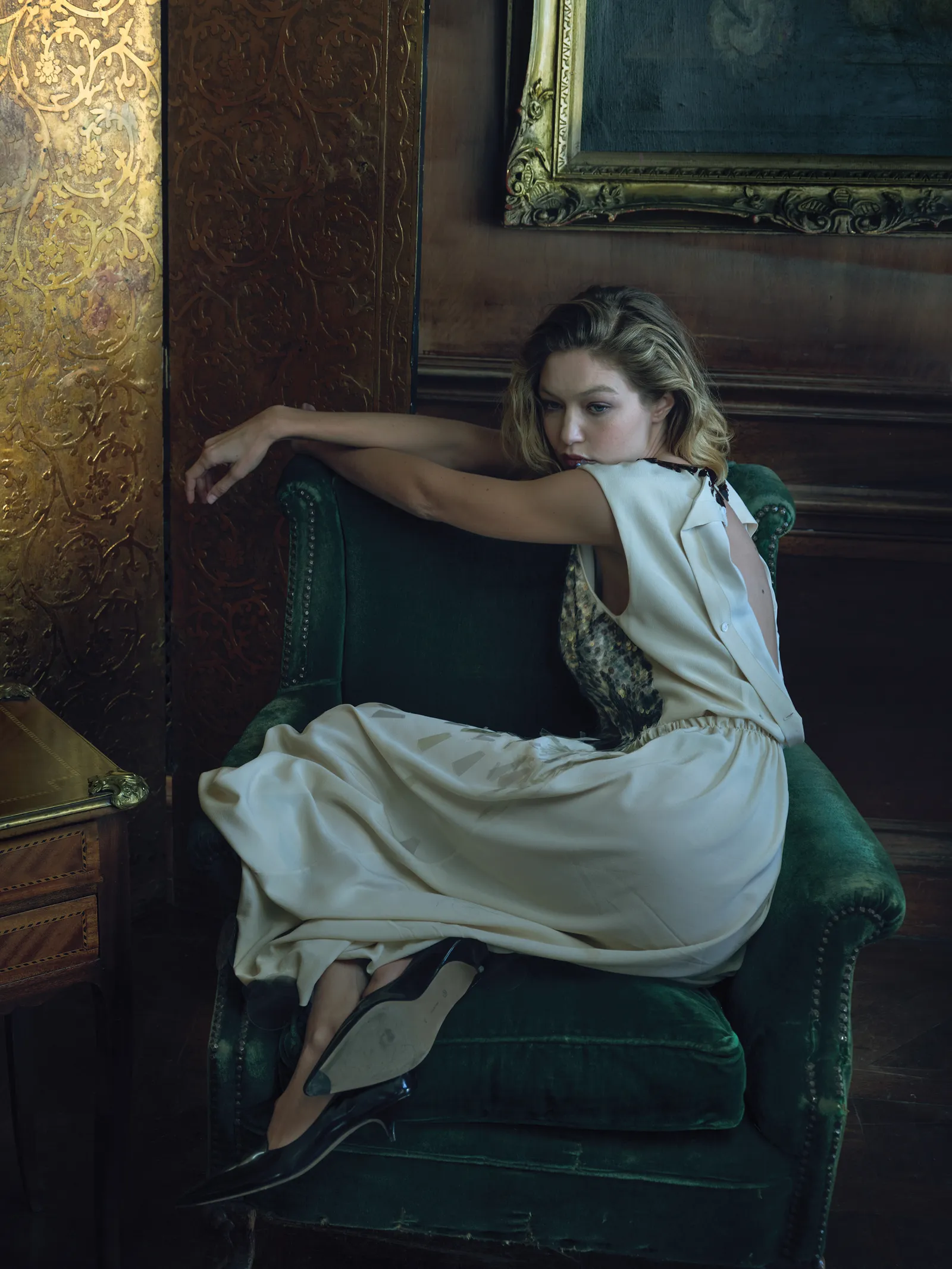 A female fashion model with blonde hair sits in a green velvet chair in an elegant room wearing a cream-colored dress and black pumps.