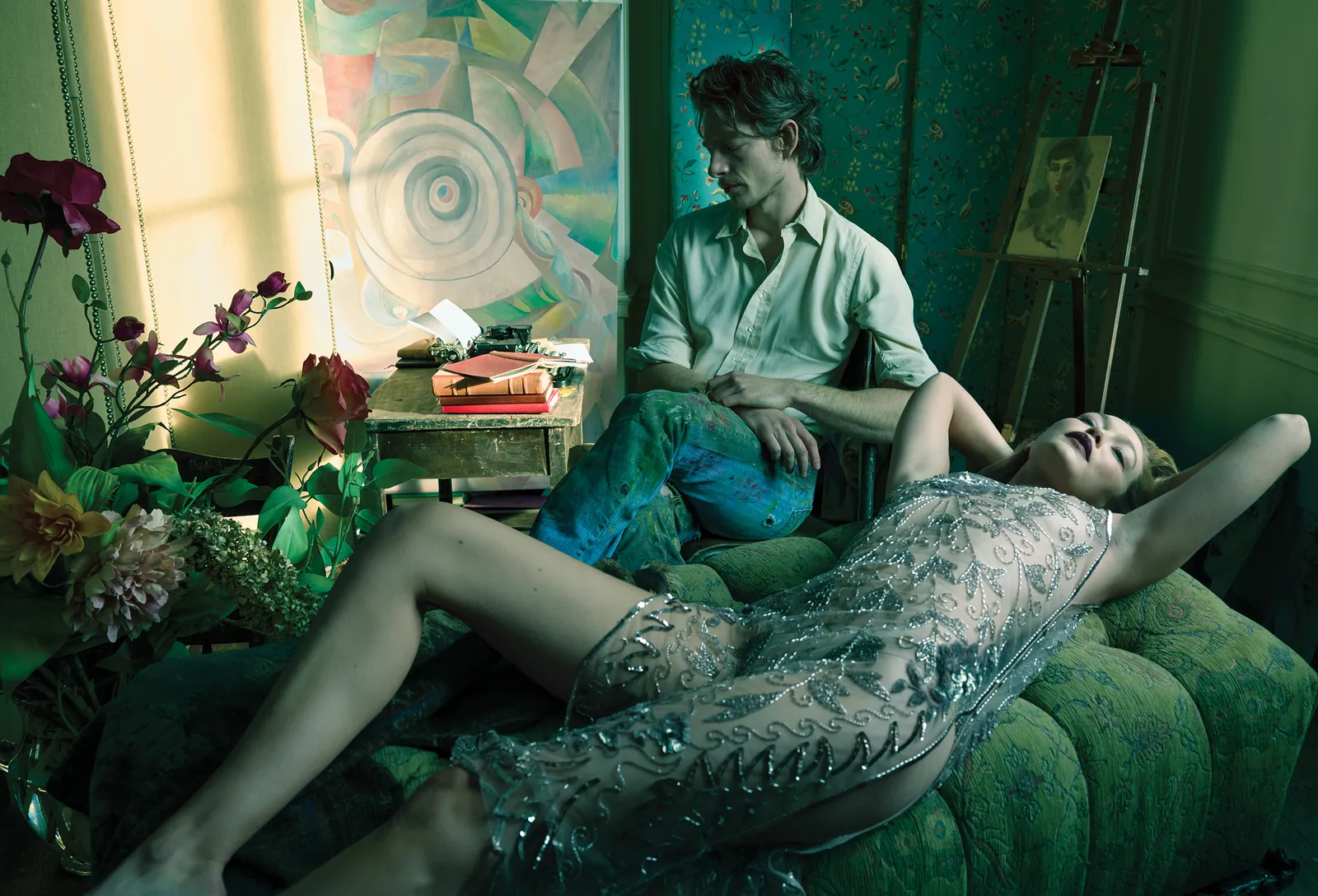 An adult male seated near an adult female reclining on a green couch, surrounded by flowers and abstract art in a brightly lit interior.