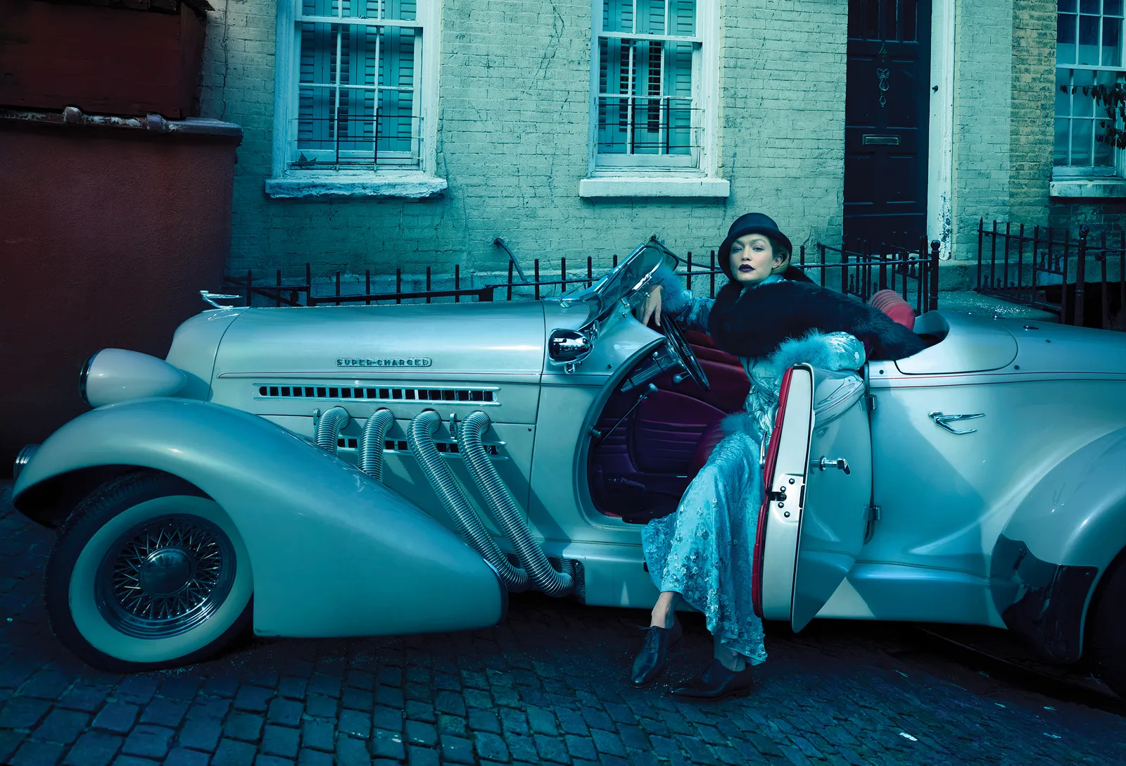 Woman in retro outfit sitting on the door of a vintage car parked on a brick street in front of a building with brick facade.